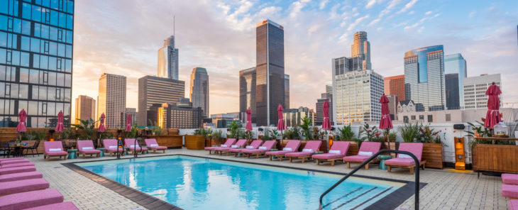 a pool with pink lounge chairs and umbrellas on a rooftop with a city skyline in the background