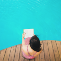 a woman sitting on a deck reading a book by a pool