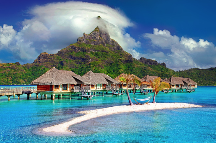a group of houses on stilts in a body of water with a hammock with Bora Bora in the background