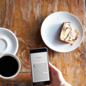 a hand holding a phone next to a plate of coffee and a piece of cake