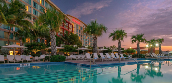 a pool with palm trees and chairs in front of a hotel